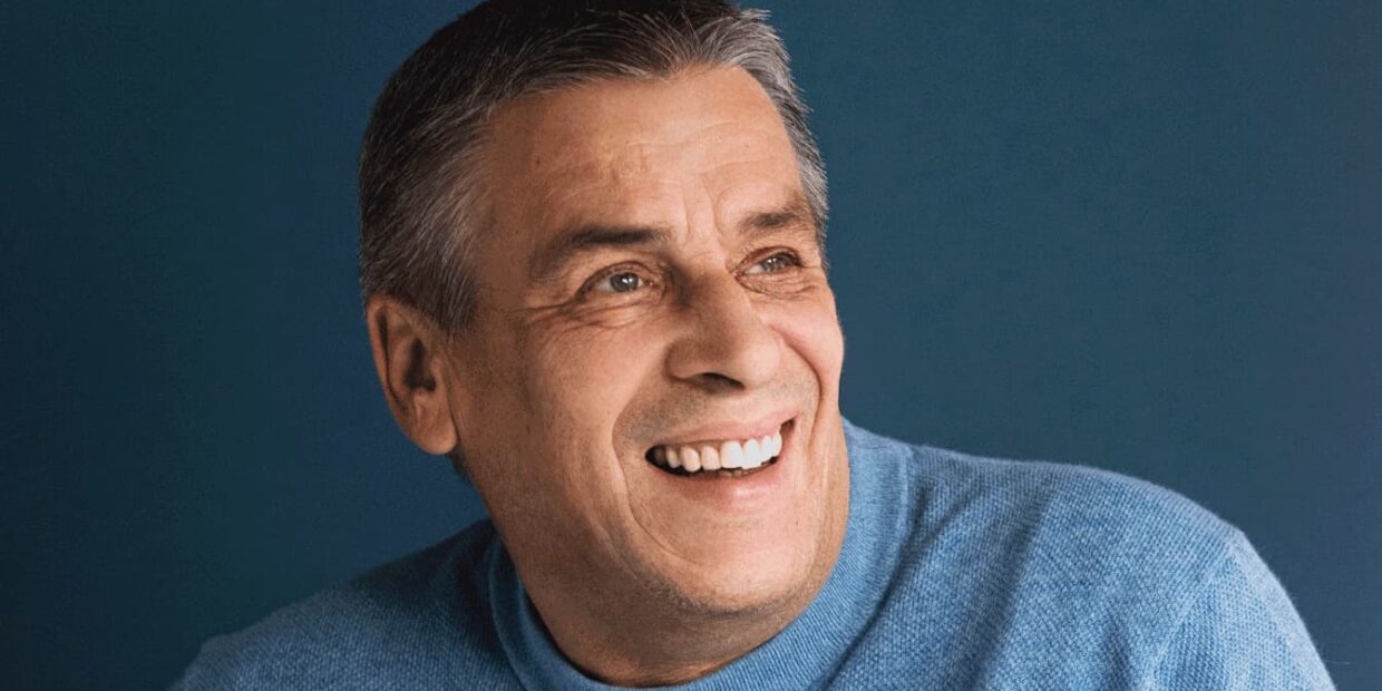 Portrait eines älteren Herrn der lächelt und einen blauen Pullover trägt.